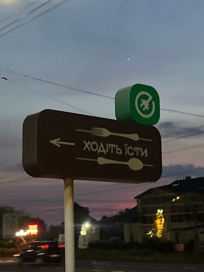 In Strumivka, Ukraine, a dark brown directional sign with a rounded edge is mounted on a light-colored pole. The sign features white text that reads "ходіть їсти" and is accompanied by stylized icons of a fork and a spoon. Above the brown sign, a green square object with a white circular logo is visible against a twilight sky. The sky is a blend of pale blue and dusky pink hues, with a thin crescent moon visible in the upper right portion. Power lines traverse the sky horizontally. In the background, a blurred building with large windows and interior lights is partially visible on the right. On the left side of the image, a red neon "TAXI" sign is also blurred. The scene appears to be near a road, with a dark vehicle in motion and blurred to the left of the sign.