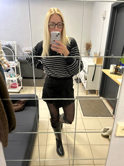 The image captures an interior scene, reflected in a segmented mirror, within a commercial establishment in Voskresensk, Russia. The primary subject is a fair-skinned female, positioned centrally, taking a selfie. She has long blonde hair, wears black-rimmed glasses, a black and white horizontally striped long-sleeved sweater, a black mini-skirt, sheer black tights, and black knee-high boots. She holds a light-colored smartphone with a multi-lens camera in her right hand, extended to capture her reflection, while her left hand rests on her hip. Dark nail polish is visible on her fingers.

In the reflected background on the left, a second individual is partially visible, wearing a light-colored top and blue jeans, standing near a white, three-tiered rolling utility cart holding various small bottles and containers, indicative of a beauty or personal care setting. Behind the cart, a dark gray sofa or bench is visible, with a brown handbag resting on it. A textured, brown, fuzzy garment is draped over a foreground object.

On the reflected background's right side, a white dresser with light wooden drawers and legs is present. On its top surface are decorative dried plant stalks in a vase, a small wooden box, and other items; one drawer is slightly open. Adjacent to this, a dark wooden countertop holds a brown bottle, a roll of white paper (possibly tissues), and artificial green foliage.

The floor is tiled with light-colored square tiles, with a dark rectangular mat visible near the dresser. A pair of light-colored, possibly fur-lined, slippers are on the floor in the lower right. The walls are light gray or white, and an electrical outlet is visible on the far right wall, alongside a dark doorframe. The room is uniformly lit by interior artificial lighting.