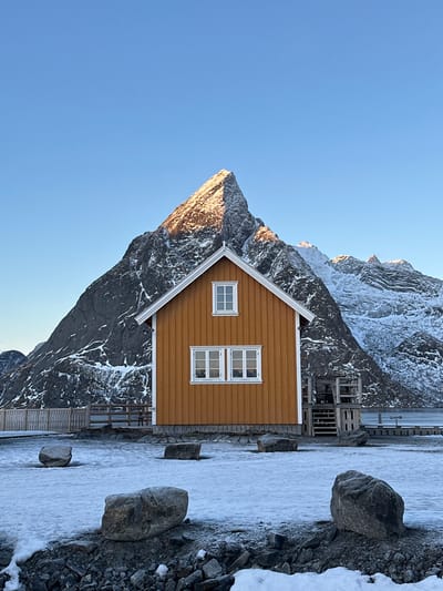 The image displays a winter landscape in Hamnøya, Norway. A single, two-story, gabled wooden house with vertical orange siding and white trim stands centrally. The house features a single white-framed window on its upper floor and two adjacent white-framed windows on its lower floor. To the right of the house, a wooden staircase and platform are visible. Immediately behind the house, a towering, jagged mountain, partially covered in snow and dark rock, rises prominently. The mountain's peak is illuminated by direct sunlight from the left, casting a warm glow. The ground in the foreground is covered by a thin layer of snow and ice, interspersed with dark gravel and several large, dark grey boulders. A wooden fence extends from the left side of the house towards the background. The sky is clear and bright blue. No individuals or animate subjects are present.