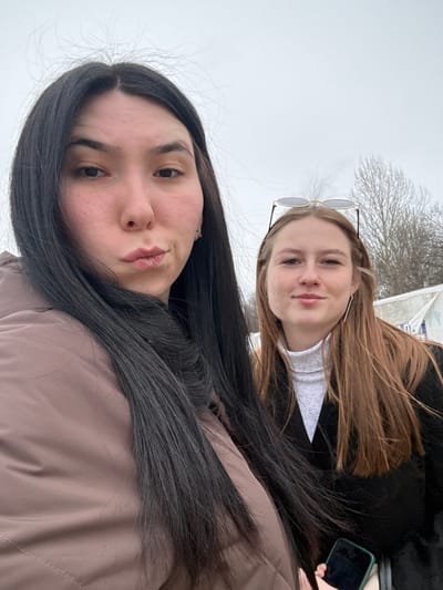 This is an outdoor selfie featuring two young women in Nizhnie Yaki, Russia, likely taken during late autumn or winter given the bare tree branches in the background. The image is oriented vertically, with the subjects appearing from a slightly downward-looking perspective.
In the foreground, a woman with long, dark, straight hair, parted down the middle, looks directly at the camera. She has a neutral expression with her lips slightly pursed and dark eyes visible. She is wearing a light brown or dusty rose-colored puffer jacket.
Behind her, partially obscured, is another young woman with long, light brown hair. She is smiling gently and also looking towards the camera. She has a pair of sunglasses pushed up onto her head and is wearing a black jacket over a white, high-necked top. Her right hand, holding a mobile phone with a dark green case, is visible on the right side of the frame.
The background consists of a bright, diffused sky, indicating an overcast day. Sparse, bare tree branches are visible against the light background on the upper left, providing a subtle environmental detail. The overall atmosphere is casual. There is no visible text within the image.