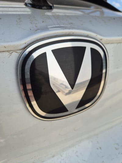 This media file is a close-up shot of a car emblem, likely a logo, mounted on a white vehicle. The emblem is oval-shaped with a metallic, reflective border and a black and silver interior design. The interior features a stylized design with sharp, angular shapes that create a sense of motion or depth. The metallic parts of the emblem reflect the surrounding environment, showing hints of a bright sky and possibly other objects.
The white surface of the car appears slightly textured and has some visible dirt or dust specks, suggesting it is not in pristine condition. The lighting indicates it is daytime, with a bright light source casting reflections on the emblem and vehicle surface.
There is no visible text in the image. The overall impression is that of a detail shot focusing on the automotive branding, with the car's exterior as the backdrop. The scene is static, highlighting the design of the emblem against a subtly weathered surface. The location context of Lenina, Russia, doesn't provide specific visual cues within the image itself.