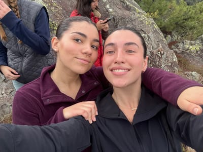 The image depicts four young adult females in a natural, rocky outdoor environment, consistent with the mountainous terrain in the vicinity of Huasca de Ocampo, Mexico. In the foreground, two females are positioned closely, appearing to take a selfie. The female on the left wears a dark purple fleece pullover, has dark hair pulled back, and a single pearl earring in her left ear; her left arm is positioned around the right female's shoulder. The female on the right, holding the camera, wears a black hooded jacket and a gold necklace with a "Roberta" pendant; her right arm is extended. Both foreground females are smiling and looking directly at the camera. In the background, on the left, a third female is partially visible from the chest up, wearing a grey quilted vest over a dark blue long-sleeved shirt, with a light-colored braid draped over her shoulder. On the right background, mostly obscured by a large rock, a fourth female in a red jacket is visible, looking down at a smartphone she holds. The environment consists of large, lichen-covered grey rock formations, sparse pine trees, and dry ground cover under an overcast sky.