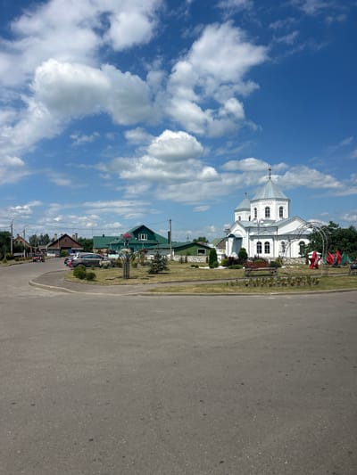 The image depicts a roadside view of a village in Belarus. The foreground is dominated by a gray asphalt road. To the left of the road, a line of houses and a white church with a silver dome are visible. The church has a cross on top and several arched windows. Some of the houses are green, while others are made of wood. Flowerbeds and greenery surround the buildings. In the background, trees and a clear blue sky with scattered white clouds create a bright, daytime scene. The orientation of the image is rotated 90 degrees counter-clockwise from standard. There are no people visible in the image.