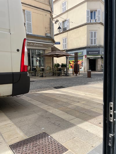 The image shows a street scene in Meaux, France. In the foreground, partially visible on the left, is the rear of a white van. The street is paved with light-colored stone tiles, with a darker cobblestone area visible further back. On the right side of the image, a dark metal frame is present.
In the mid-ground, there is an outdoor seating area for a brasserie, with several round tables and chairs set up under a large umbrella. The brasserie is identified by a sign reading "De La Cathédrale". Adjacent to the brasserie is a "Rapid Market" store with a sign that says "Bienvenue".
In the background, buildings line the street, featuring typical French architecture with shutters on the windows. A street sign labeled "RUE SAINT REMY" is visible. The scene appears to be a pleasant, sunny day.