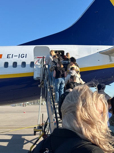 The image depicts passengers boarding a commercial aircraft at an airport, likely in Weeze, Germany. The perspective is from behind a person with long blonde hair, who is in the immediate foreground, looking forward towards the aircraft. A line of other passengers is visible ahead, ascending a mobile boarding staircase to enter the aircraft's fuselage.
The aircraft is a distinctive dark blue and white, separated by a yellow stripe running horizontally along its side. The registration number "EI-IGI" is clearly visible in large black letters on the white part of the fuselage, identifying it as a Ryanair Boeing 737. The boarding stairs are metal, connecting the tarmac to the aircraft door.
The scene takes place under bright, clear blue skies, indicating good weather conditions during the daytime. The tarmac can be seen below the aircraft and stairs, with a small orange cone faintly visible in the distance on the right side of the tarmac. The overall impression is a routine and busy aircraft boarding process.