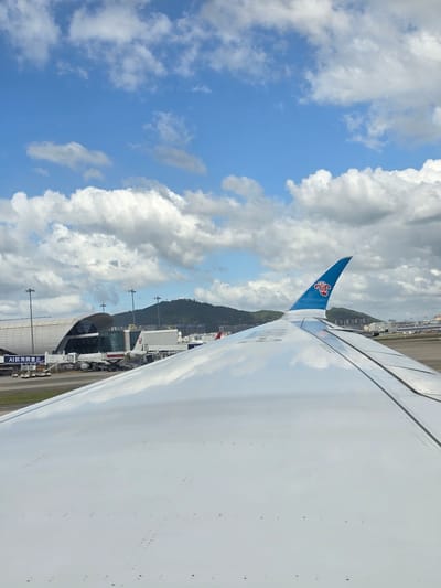 The image is taken from the perspective of an airplane window, looking out at an airport tarmac. The foreground is dominated by the reflective white surface of an airplane wing, with visible rivets and seams. A blue and red logo, the China Southern Airlines logo, is visible on the wingtip. In the middle ground, several airplanes are parked at gates connected to a modern airport terminal with a distinctive curved roof. Various airport service vehicles and equipment are present on the tarmac. In the background, lush green hills rise behind the airport buildings. The sky above is a vibrant blue, dotted with fluffy white clouds. The scene appears to be at Bao'an District, China, likely Shenzhen Bao'an International Airport.