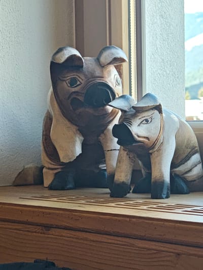 The image shows two wooden pig figurines on a wooden shelf in front of a window. The larger pig is on the left, and a smaller pig is on the right. Both pigs are carved in a stylized manner with distinct facial features, including prominent ears and snouts. The larger pig appears to have a darker, wood-grain finish, while the smaller pig is lighter with darker accents. The background shows a textured wall on the left and a window on the right, through which a blurred landscape of mountains and trees can be seen. The shelf itself is made of natural wood with visible grain and texture. The lighting suggests it is daytime, with light coming from the window. The overall setting appears to be indoors, possibly in a residential or decorative space. The specific location in Filzen, Austria, is not explicitly evident from the image itself, but the context suggests a potential placement within a home or establishment in that area.
