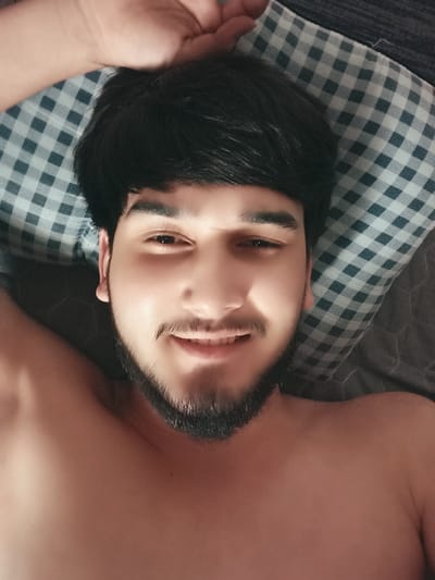 A man with dark hair and a beard is lying on a pillow, looking at the camera. His left arm is resting on his head, and his chest is visible. The pillow has a checkered pattern of white and blue. The background is dark and appears to be a bed or sofa. The image was taken in Hongshan District, China.