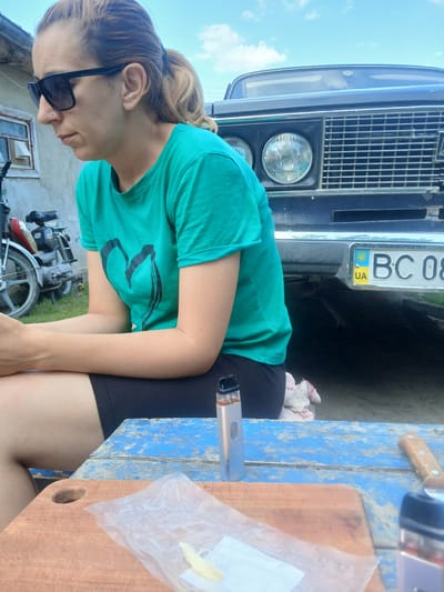 The image shows a woman sitting at a rustic blue wooden table outdoors. She is wearing sunglasses and a teal t-shirt with a black heart design. In front of her, on the table, are a cutting board with a plastic bag containing a yellow piece of food, a vape pen, and a knife. Behind her, a dark-colored car with a Ukrainian license plate (BC 08 UA) is parked. A motorcycle is also visible in the background, to the left of the woman. The setting appears to be a residential area, possibly in the city of Pishchanka, Ukraine, given the Ukrainian license plate. The sky is blue with scattered clouds.