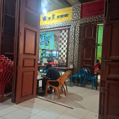 This image captures the interior of a cafe in Rhieng Blang, Indonesia. The scene is viewed through a doorway, with a person hunched over a table in the foreground, appearing to be working or preparing something. Orange and blue plastic chairs are visible, along with stacked red chairs in the lower right corner, suggesting a busy or soon-to-be-busy establishment.

The cafe's walls are decorated with a stone-patterned tile and a bright yellow mural featuring a teapot and cups, along with the word "Coffee" in large, stylized blue letters. Further into the cafe, a checkered wall pattern and a cooler stocked with drinks are visible. Two other individuals are seated at a table in the background, engaged in conversation.

A red banner with the word "WELCOME" in capital letters hangs on the left side of the entrance. The overall atmosphere appears casual and lively, likely during operating hours. The lighting suggests it is daytime, though the specific time is not discernible. The tiled floor and wooden doors are prominent features of the cafe's interior design.