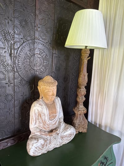 The scene is an interior arrangement in Tibubiyu, Indonesia. A wooden statue of Buddha, with a natural wood and distressed white finish, sits in a meditative posture on a dark green table. The Buddha features a textured ushnisha and hands clasped in Dhyana Mudra. To the right and slightly behind the Buddha, a tall, ornate wooden lamp with a carved, light brown base and a pleated, off-white conical shade stands on the same green table. The table itself is painted dark green with a slight curve visible on its right side, featuring faint decorative accents. In the background, a dark wooden wall or panel is intricately carved with repeating patterns of concentric circles, sunbursts, and geometric shapes. To the far right, white, vertically pleated curtains are visible, suggesting a window or opening. The objects are static, arranged for display.