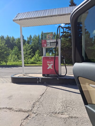 A gas pump at a station is depicted under a white awning in Glāžšķūnis, Latvia. The pump is red and white with the "Multi X" logo prominently displayed. A fuel hose hangs from the pump, ready for use. The surrounding area is paved with a textured concrete surface, which shows signs of wear and cracking. Green trees and lush vegetation form the background, under a clear blue sky. The right side of the image shows a partial view of a car, including its door and window, suggesting the photo was taken from inside a vehicle.