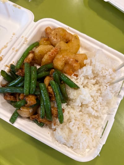 Here is a description of the image:

The image shows a white takeout container filled with food. The container holds white rice, fried shrimp, and green beans stir-fried with meat. Chopsticks rest atop the rice. The container sits on a light green surface. The location could be Honolulu, United States of America.