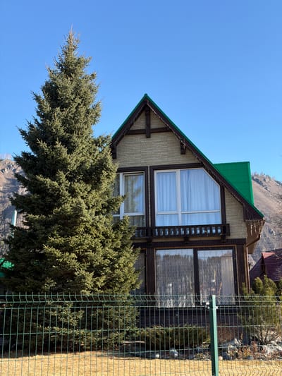 The image shows a two-story building with a green roof located in Manzherok, Russia. The building has a triangular upper portion with two windows. A large evergreen tree partially obscures the lower portion of the building. A green metal fence is visible on the right side of the image. The sky is clear and blue. The terrain in the background is mountainous.