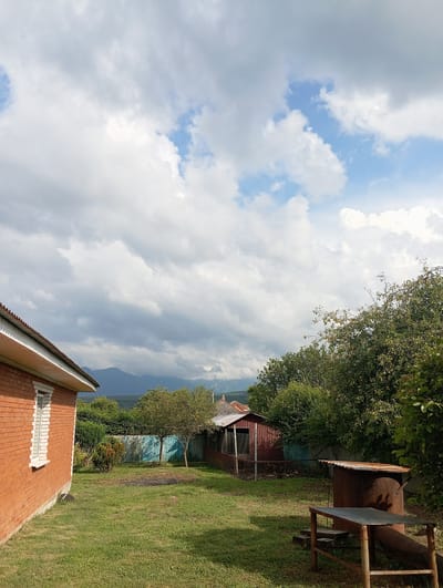 The image depicts a residential backyard in Chernigovskoye, Russia, characterized by a brick house on the left, a lawn, a red corrugated metal shed, and a rusty metal structure resembling a makeshift grill or brazier in the foreground. The yard is surrounded by a fence and mature trees, with mountains visible in the distant background under a partly cloudy sky. The scene is set during daylight hours.