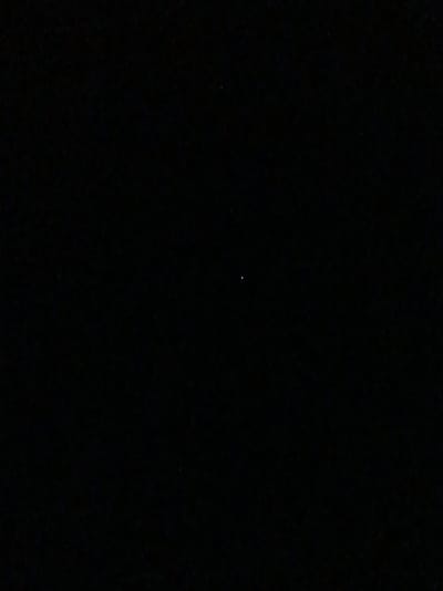 The image is a dimly lit, dark scene, likely outdoors at night, possibly in a rural setting. The most prominent feature is a single, small, bright white point of light located slightly to the left of the vertical center and about a third of the way down from the top. This light source is likely a star, planet, or a distant artificial light. The rest of the image is obscured by darkness, with very faint, scattered speckles of red and blue light visible, which could be distant lights or sensor noise. There are no people, discernible objects, or recognizable landmarks visible due to the extreme darkness and low light conditions. The location in Шарпілаўскі сельскі Савет, Belarus, suggests a potentially rural or semi-rural environment, which aligns with the dark, unpopulated appearance of the image.