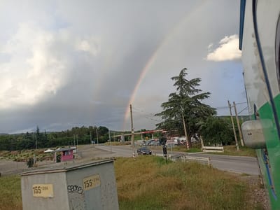 The image depicts a street scene in Marino Pole, Bulgaria, characterized by a prominent double rainbow arching across a cloudy sky. In the foreground, a gray utility box with yellow numerical stickers is visible. To the right, a portion of a green and white vehicle is in frame. The middle ground features a road with a black car driving on it, a gas station, and several parked vehicles. Lush green trees and vegetation are present throughout the scene, with a tall evergreen tree on the right. A pink booth or kiosk is situated in the distance, surrounded by plants. The overall atmosphere is that of a potentially rainy or recently rained-upon day.