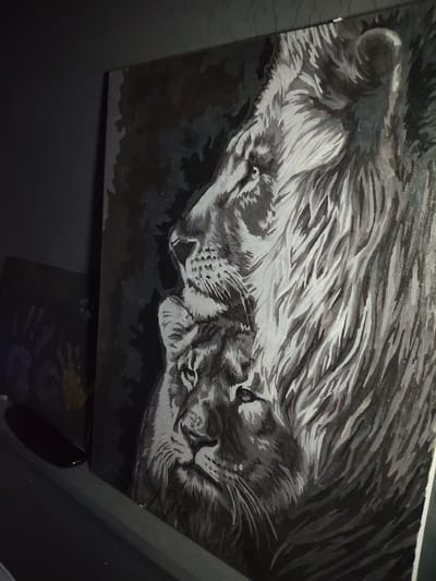 The image shows a painting of two lions, a male and a female, in a monochromatic style with shades of black, white, and grey. The male lion is larger and positioned behind and slightly above the female, with its mane prominently featured. The female lion is in the foreground, looking towards the left. The background is dark and abstract, with visible brushstrokes. The painting is displayed on a wall, and to the left of the painting, another artwork with handprints is partially visible. The scene appears to be indoors, with low lighting. No specific location in Reftinsky, Russia is discernible from the image itself.