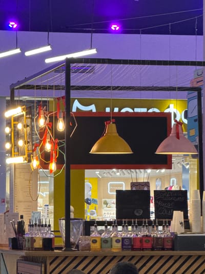 This image captures an interior view of a commercial establishment, likely a cafe or bar, characterized by an eclectic lighting design and a service counter. The scene is brightly lit by various artificial light sources, suggesting an indoor setting or nighttime.
In the foreground, a decorative metal framework supports multiple light fixtures, including horizontal fluorescent tubes and an elaborate arrangement of glowing Edison-style bulbs with red wiring. A prominent feature in the midground is a large black rectangular sign with a red border, in front of which hang two bell-shaped pendant lights, one pink and one yellow.
To the right, a counter displays an array of bottles, many fitted with pour spouts, along with small colorful boxes (possibly tea or coffee), and stacked paper cups. Two small black chalkboards rest on the counter. The upper chalkboard reads "Тортик Каппучино" (Cake Cappuccino), while the lower one has "Соус" (Sauce). A vertical white sign behind the bottles partially shows the word "Счастье" (Happiness).
The background features brightly lit yellow and light blue wall panels, with some indistinct white Cyrillic text visible. On the far right, a section of decorative wooden paneling with horizontal stripes adds texture. On the far left, bright purple LED lights are visible. A dark, indistinct shape on the far right edge of the frame might be a person's head. The overall atmosphere is vibrant and functional, typical of a modern food or beverage service area.
