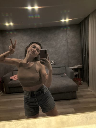 An indoor mirror selfie captures a young woman posing in what appears to be a living room or bedroom. The image is oriented with the subject lying horizontally, indicating it was likely taken with the phone held vertically and then the photo rotated for viewing.
The woman, with dark hair, is visible from approximately the chest down to the mid-thigh. She is wearing a fitted light brown or beige athletic crop top and light blue denim shorts. Her left arm is extended towards the camera, holding a dark-colored smartphone, possibly purple or dark red, with two visible camera lenses. Her right arm is bent at the elbow, and her hand is held up, displaying a "peace" sign (V-sign) with her index and middle fingers extended. She has a slight smile on her face. A small, dark tattoo or mark is visible on her left upper arm.
The background, visible through the mirror's reflection, shows a room with a somewhat dim and warm ambiance. On the left side of the reflection, bright, multi-colored light flares and streaks are prominent, suggesting reflections from a light source or window, possibly creating diffraction effects due to the mirror surface or camera lens. Beyond the subject, in the reflected space, there is a dark grey, textured wall and a grey sofa or couch with a subtle pattern. On or near the sofa, various items can be vaguely discerned, including what looks like a pair of white sneakers and possibly a light-colored blanket or clothing item. The floor, visible at the bottom of the reflected image (right side of the actual room), is made of light-colored wooden planks. The mirror surface itself appears to have some dust or minor imperfections.