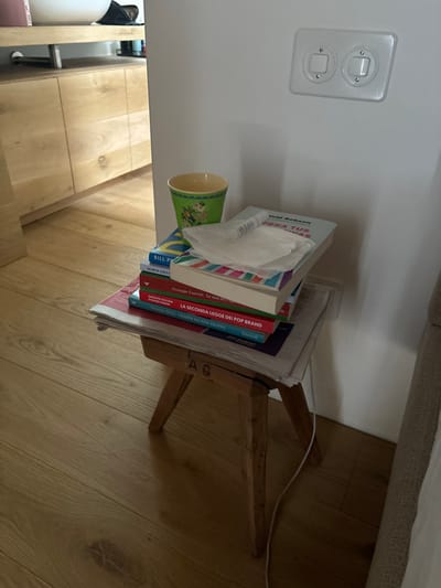 The image shows a wooden stool with four legs, located in a room with light wood floors and white walls. The stool is near a corner where a white wall meets a beige upholstered wall. On the stool, there are five books stacked in a pile. On top of the books is a white napkin, and a green cup with a yellow rim sits on top of the napkin. The books are all in Italian and the titles are visible. The book on top is titled "PERA TUA". The other book titles include "LA SECONDA LEGGE DEI POP BRAND" and "BILL P". There are also a couple of newspapers on the stool. The white wall has a single light switch plate with two switches. The room is in Celerina/Schlarigna, Switzerland, with the coordinates 46.514, 9.859.