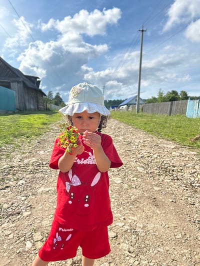 In Новомуллакаево, Russia, on a sunlit day with scattered clouds, a young girl stands on a gravel road. She wears a red t-shirt and red shorts, both adorned with bunny graphics. On her head, she sports a pale green hat with lace trim and floral embellishments. In her hands, she holds a small bunch of wild strawberries. The road is flanked by grassy areas and wooden fences. In the background, wooden houses and a utility pole are visible.