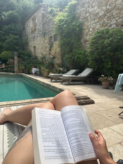 A person is sitting near a swimming pool in Saint-Victor-la-Coste, France, reading a book with Hebrew text. The person is fair-skinned, wearing striped fabric as their seat. Their right foot is visible in the lower right corner. The book occupies the right portion of the frame, obscuring the person's upper body and providing a partial view of their right hand holding the book open. The swimming pool extends from the lower center to the middle left of the frame. In the background, there's an old building covered with greenery, suggesting a historic architectural style. Two lounge chairs are visible near the pool, further back and to the left.