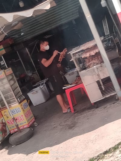 A man wearing a white face mask and a dark t-shirt stands in front of a food stall in Ben Cau, Vietnam. He is holding a skewer of grilled meat in his right hand and a small knife or pick in his left. The food stall features a metal grill with more skewers of meat cooking, a red stool, and various containers and signs. A yellow sign with Vietnamese text is visible on the left. The ground is paved, and a large tire is positioned near the stall. The background suggests an outdoor market or street scene with structures and awnings.