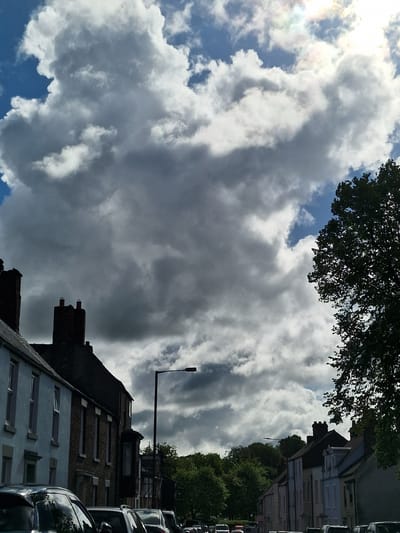 The image shows a view of a cloudy sky above a residential street. The sky is filled with large, gray and white cumulonimbus clouds, with patches of blue sky visible in the lower left corner. In the upper center of the frame, the silhouette of a tree's foliage is visible.
Below the sky, the tops of buildings can be seen. These are traditional brick buildings with pitched roofs and chimney stacks. A street lamp extends from the building. Cars are parked along the side of the street.
The location is likely Durham, United Kingdom, based on the architectural style of the buildings and the general environment.