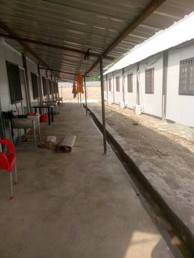 The image depicts an outdoor scene in Indo, Nigeria, likely a compound or workers' quarters. The foreground shows a concrete walkway extending to the left, leading towards a series of modular buildings constructed with corrugated metal walls and roofs. These buildings appear to be single-story units, each fitted with barred windows and air conditioning units on the exterior.
On the walkway, to the left, are several metal sinks with plumbing, a red plastic chair, and various items like boxes and rolled-up materials. A bright orange tarp or piece of fabric is visible hanging from a support structure above the walkway, along with what appears to be a black object clipped to a wire. Further down the walkway, more sinks, tables, and orange tarps are visible.
The buildings are situated alongside a narrow, partially filled drainage channel that runs along the edge of the walkway. The area between the drainage channel and the buildings is a dirt and gravel path. The background reveals a brick wall and some vegetation, suggesting the compound is in a semi-urban or rural setting within Indo. The overall impression is one of a functional, possibly temporary, living or working space.