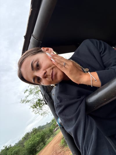 A woman in a black shirt poses in what appears to be an off-road vehicle. She has brown hair, brown eyes, and long white nails. She wears a gold chain necklace, a gold hoop earring, a ring, and a white bracelet. She is holding her hand to her face. The sky is overcast. The vehicle is surrounded by trees and a dirt road.