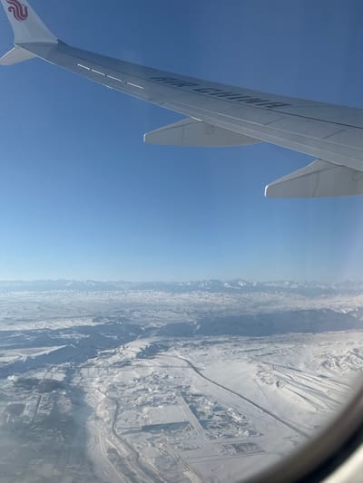 An aerial view from an aircraft window depicts a vast, snow-covered landscape under a clear, bright blue sky. The upper left portion of the image shows the white wing of an Air China aircraft, emblazoned with the "AIR CHINA" text and the phoenix logo on the wingtip.

Below the aircraft wing, the ground stretches into the distance, characterized by undulating, extensively snow-covered terrain. A prominent range of snow-capped mountains forms the distant horizon. In the midground and foreground, a significant highway or major road, dark in contrast to the snow, curves through the landscape from the left towards the lower right. Adjacent to this road and across the snow-covered plain, multiple large, rectilinear complexes are visible. These complexes appear to be industrial parks, logistical centers, or development zones, featuring numerous buildings and structured layouts. The scene is consistent with an overview of the Ürümqi Economic and Technological Development Zone (ETDZ) or other industrial areas situated to the south-southeast of Ürümqi Diwopu International Airport (URC) in Ürümqi, Xinjiang Uyghur Autonomous Region, China, during winter conditions. The environment is clear, indicating excellent visibility on a cold day. No individuals are discernible on the ground.