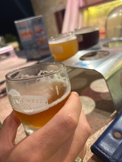 A close-up perspective depicts a light-skinned hand, exhibiting a metallic ring on the left ring finger, grasping a clear glass. The glass, approximately half-filled with a light amber liquid consistent with beer, shows residual foam and condensation. The visible branding on the glass reads "EAST WEST COMPANY EST. 2014". The hand is positioned over a light-colored, textured table surface featuring faint circular patterns. Adjacent to the hand, a dark blue leather wallet is partially visible. In the midground, a metallic flight tray holds two smaller glasses; one contains a golden-yellow liquid, and the other a dark reddish-brown liquid. A blurry, illuminated vertical menu board displaying colorful graphics is discernible in the background. The environment is dimly lit. This scene is located at an East West Brewing Company establishment in An Hải Ward, Da Nang, Vietnam.