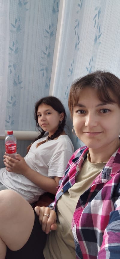 Two females are seated. The female in the foreground is on the right, facing the camera and smiling. She has brown hair styled with bangs and is wearing a tan t-shirt with a pink and blue plaid shirt unbuttoned over it. She has a gold hoop earring in her visible ear and a ring on her left index finger. The female in the background is on the left, looking towards the camera with a neutral expression. She has dark brown hair in two braids and is wearing a white t-shirt. She is holding a red bottle of water. The background appears to be a room with light blue curtains that have a leaf pattern. There is no identifiable information about the city of Tashkazgan, Russia in the image.