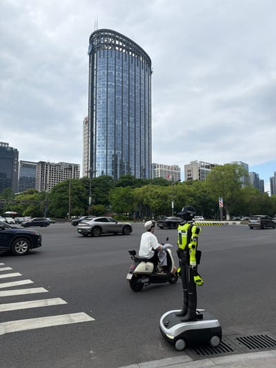 The image depicts a street scene in the Binjiang District of China, featuring a tall, curved skyscraper. A police officer is standing next to a black and white autonomous driving base (robotaxi), positioned near a street drain. The officer is wearing a helmet, a high-visibility yellow and black jacket, and black pants. An individual, possibly a passenger, is seated on a scooter alongside the robotaxi. The street has a crosswalk with white lines. Several cars are parked along the street, including a silver car. The background includes various other buildings and trees. The sky is overcast.