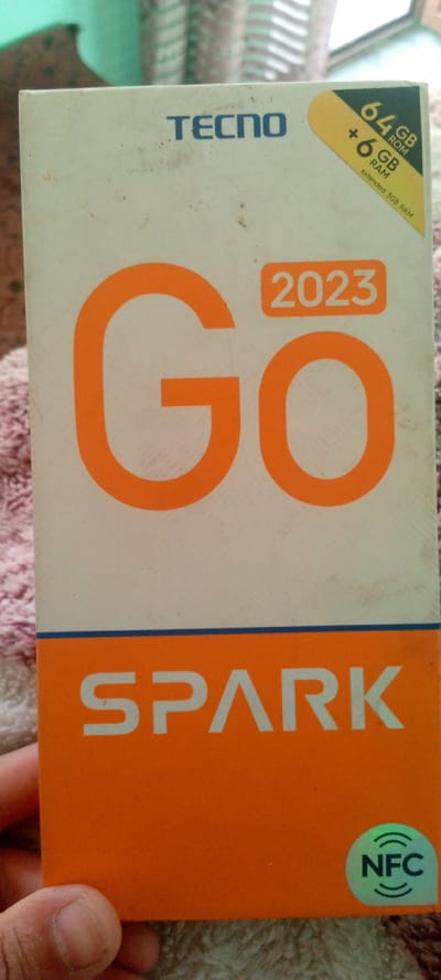 The image shows the front of a TECNO SPARK GO 2023 mobile phone box. The box is primarily off-white with large orange lettering that reads "GO" and the year "2023" in a smaller orange rectangle. Below this, in a large orange section, the word "SPARK" is written in white letters. In the top right corner, there is a yellow sticker indicating "64 GB ROM + 6 GB RAM" and "extended 3GB RAM". At the bottom right, there is a holographic "NFC" logo. The box appears slightly dirty. A hand is visible at the bottom left, holding the box. The background is blurred, showing textured fabric, possibly a carpet or blanket, and hints of other objects. The image is not taken in Marhamat, Uzbekistan.