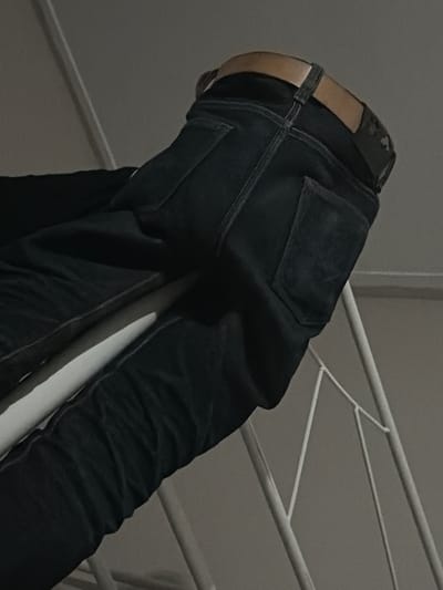 A low-angle, close-up shot depicts the rear view of a person wearing dark denim jeans and a brown leather belt. The person's legs and hips are visible, and they appear to be leaning or resting on a white metal railing. The jeans have a single back pocket visible on the right side, and the belt buckle is also partially obscured. The background consists of a plain, light-colored wall and the white metal structure. The scene is dimly lit, suggesting an indoor environment. No specific location in Tupah, Indonesia can be determined from the image.