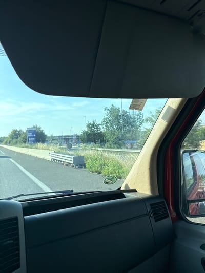 The image, taken from inside a vehicle, shows a view of a highway and a service station in Avignonet-Lauragais, France. The foreground is dominated by the vehicle's dashboard and sun visor. Outside the windshield, a highway lane marked with white lines is visible, bordered by a concrete barrier and a grassy verge. Beyond the verge, a service station with fuel pumps and a building is apparent. Trees and power lines are scattered across the landscape. The sky is blue with scattered clouds. A red vehicle is visible in the side mirror. A circular green sticker with the word "DEKRA" is affixed to the lower left of the windshield.