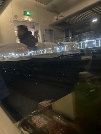 The image is a nighttime view inside a train car. A man in a dark jacket and white shirt is visible, likely using a restroom. The walls are white with various signs, including a green exit light and symbols. A window reflects the outside, showing a building at night. Dark, patterned seats are visible. Packaged food items rest on a table near the window, adding clutter to the reflection. The image has a blurred, layered effect due to the reflections.