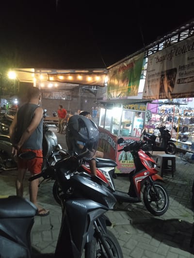 This nighttime image captures a lively street scene in Cangkring, Indonesia. The foreground features several motorcycles, with one red scooter and a black scooter prominently displayed. One man, wearing a dark grey tank top and red shorts, stands with his hands behind his back, facing away from the camera. Another person, wearing a red shirt, is visible in the background, walking away from a food stall.
The setting appears to be an outdoor market or food court, with several stalls illuminated by strings of warm lights. A stall with the word "SEMPOLAN" in yellow lettering on a sign is visible, suggesting it sells a local snack. A banner with text, partially obscured, hangs above the stalls. One banner reads "BAYU AJI Collect" and another has "Artoman" written on it. The ground is paved with interlocking bricks. The atmosphere seems casual and bustling, typical of a night market.
