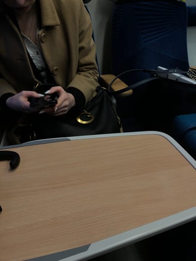 The image shows a person seated on a train or airplane, wearing a brown coat and holding a cell phone with white nails. A black purse rests on the person's lap. A wooden tray table with a white border is positioned in front of the person. The seat is blue. The lighting is soft.