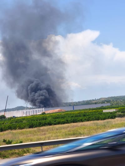 The image depicts a large plume of black smoke emanating from a building under construction in Riba-roja de Túria, Spain. The smoke is billowing into a bright blue sky with scattered white clouds. In the foreground, a highway barrier and the blurred reflection of a vehicle's interior are visible, suggesting the photo was taken from a moving car.

The construction site appears to be a large industrial or commercial building, with a white structural frame and a pile of earth or debris. Two cranes are present at the site, one tall and slender, and another shorter and thicker. In the middle ground, a green field of vegetation, possibly a vineyard or orchard, stretches across the landscape. Beyond the construction site and fields, rolling hills covered in trees and vegetation form the background. A power line tower is also visible in the distance. The overall scene suggests a fire or explosion has occurred at the construction site.