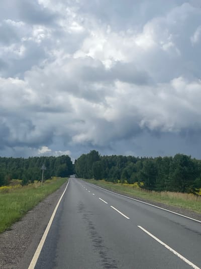 The image depicts a rural road stretching into the distance, flanked by dense green trees on either side. The sky above is filled with heavy, dark clouds, suggesting an overcast or impending stormy weather. The road itself is asphalt with white lane markings. There are no people or vehicles visible in the frame. The environment appears to be a natural landscape, likely in a rural area.