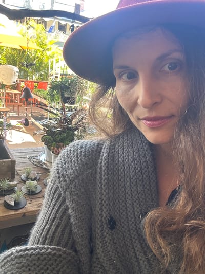 The image is a close-up, slightly angled selfie of a woman wearing a burgundy hat and a grey knitted cardigan. She has long brown hair and is looking towards the camera with a neutral expression. The background appears to be an outdoor patio or garden area, with plants, tables, chairs, and a yellow umbrella visible. Sunlight is casting shadows and highlights throughout the scene. There are no discernible landmarks that would place this location specifically in Bellingen, Australia.