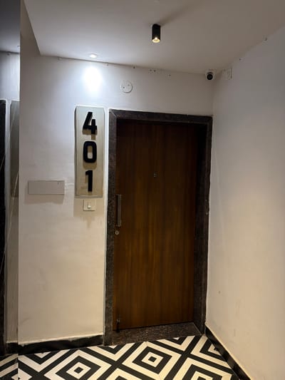 The image shows a doorway with the number "401" displayed vertically on a light-colored plaque to the left of the door. The door itself is made of dark wood with a small, circular peephole and a silver handle. The doorframe has a dark, speckled stone trim. A light switch is visible below the number plaque. The walls are painted white. Above the door, a security camera is mounted on the ceiling in the corner. The floor is tiled with a bold black and white geometric pattern. The scene appears to be in an indoor hallway, likely in Shamshabad, India, given the context provided.