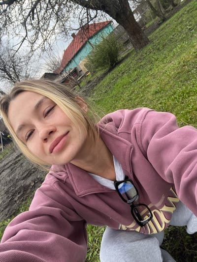 The image is an upward-angled selfie taken outdoors, likely by the subject herself. The person is a young woman with blonde hair and light skin. She is wearing a pink sweatshirt with a gold and brown design and a white undershirt. Black-framed glasses are resting on her sweatshirt near her neck. She is sitting down, possibly on the ground, as indicated by the grass and a tree trunk in the background. A building with a red roof and teal siding can be seen in the distance, along with bare tree branches against a cloudy sky. Based on the presence of a building with a traditional design and the surrounding landscape, the location might be a rural or suburban area in Bobrov, Russia.