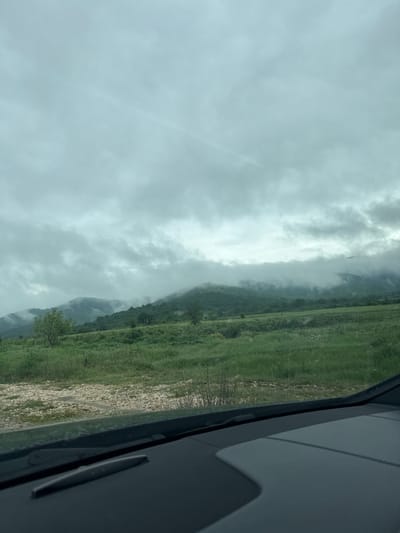 The image shows a view from inside a vehicle, likely a car. Outside the window, there is a landscape with a sky filled with gray clouds. A mountain partially obscured by mist is visible. The ground near the mountain is grassy, with some rocky areas closer to the viewer. This may be the edge of the city of Kliment in Bulgaria.