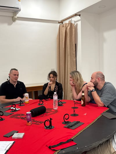 Four people, two men and two women, are seated around a round table covered with a red cloth. The table is adorned with black decorative elements that resemble numbers or letters, and also holds various items, including cans of beverages, a water bottle, a Bluetooth speaker, and a cell phone. The individuals appear to be engaged in conversation or listening attentively. The background reveals a white wall, an air conditioning unit mounted high on the wall, and beige curtains hanging on the right side of the room. The setting suggests an indoor gathering or meeting. The image was taken in Ban Rong Ruea Kham, Thailand.