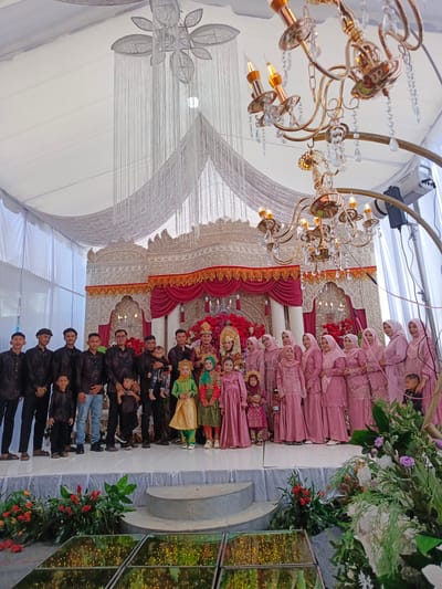 The image captures a wedding ceremony scene, likely held in a decorated tent or hall in Meuliek, Indonesia. A wedding party is posing for a photograph on a raised platform. In the center, a bride, adorned in traditional attire and a golden headpiece, is seated. She is surrounded by a group of women, all wearing matching pink traditional dresses and headscarves. To the left of the bride, a groom, also in traditional attire, stands with a group of men. Several children, dressed in traditional outfits, are interspersed within the group. The backdrop features an ornate stage with red drapes and intricate white carvings. Chandeliers hang from the ceiling, adding to the festive ambiance. In the foreground, floral arrangements and a mirrored surface with lights create a decorative element. The overall setting suggests a formal and celebratory occasion.