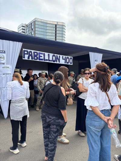 The image captures an outdoor public scene during daylight, likely an official event or polling station, set against the backdrop of modern city buildings in Santiago de Surco, Peru.
In the foreground and midground, a crowd of approximately twenty individuals, predominantly adults, is gathered. On the far left, a woman with auburn hair tied back, wearing a white long-sleeved shirt, a light grey wrap-around skirt, black pants, and black and white sneakers, stands with her back to the viewer, intently examining a white sheet of paper affixed to a temporary white fabric panel. This sheet displays text, including "N° DE MESA 051268" and a partial name "PRADO GUEVARA CARLA STEPHANIE".
Immediately to the right of this woman, another woman with her back to the camera wears a dark t-shirt and dark grey camouflage-patterned pants. Further right, a woman in a white loose-fitting top with ruffled sleeves, light blue wide-leg jeans, and a light brown visor on her head, also faces right, holding a clear plastic water bottle with a blue cap behind her back.
In the midground, several individuals are visible:
* A woman wearing a white t-shirt with "Calvin Klein" text, dark sunglasses, and a dark skirt, carries a black handbag and looks towards the right.
* A man in a black long-sleeved shirt holds a smartphone to his ear or face, appearing to be in conversation or taking a picture.
* Multiple individuals in military camouflage uniforms, consistent with Peruvian military attire, are interspersed within the crowd. At least two are clearly visible; one on the mid-left carries a dark rifle with a visible magazine, and another further back to the right also holds a rifle. They appear to be maintaining security or order.
* Other civilians in casual attire, including t-shirts, pants, and dresses, are standing, interacting, or observing.
The scene is dominated by a temporary outdoor structure with a dark blue upper frame and white fabric panels forming walls or partitions. A prominent white banner stretched across the structure reads "PABELLÓN 400" in large blue lettering. A smaller, partially visible sign to the left shows "ULA" and "(402)". The ground is paved, resembling asphalt.
In the background, under a cloudy sky, several multi-story modern buildings with extensive glass facades are visible, reflecting the ambient light. The architecture suggests a contemporary urban environment.
